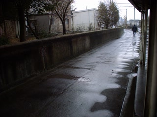 駅へ続く濡れた舗装路(移動の途中)。背景に人物が一人写る