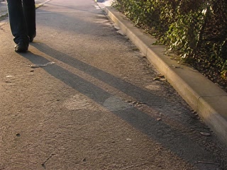 影の伸びた歩道と靴先(移動の途中)。背景に人物が一人写る