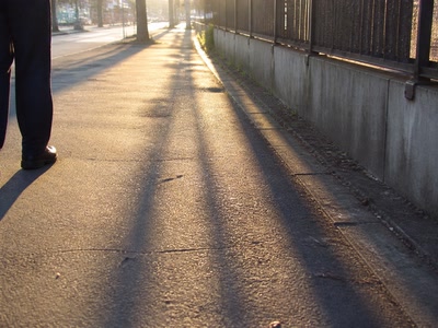 影の伸びた歩道と靴先（少し足を止めた時）。背景に人物が一人写る
