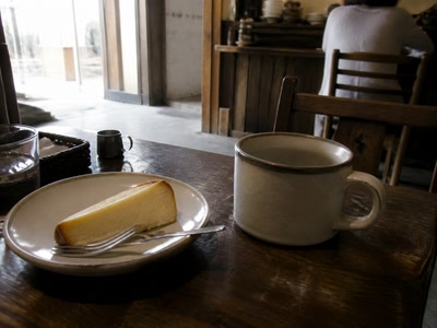 チーズケーキと厚いカップ(椅子へ座った後)。背景に人物が一人写る
