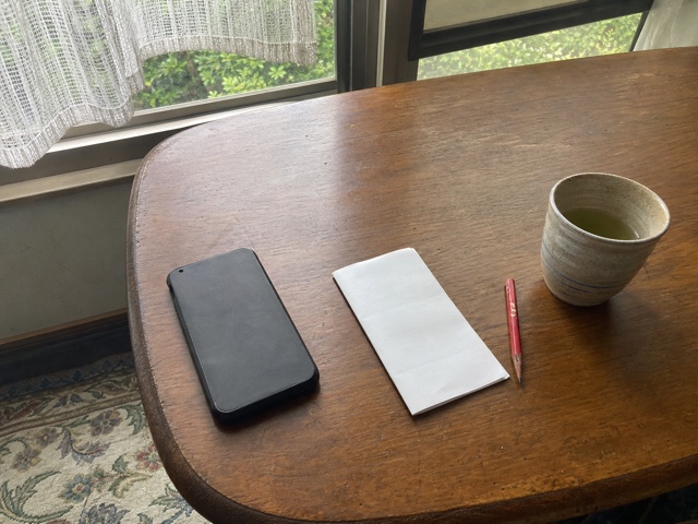窓辺の机に伏せた携帯電話と白い紙と赤鉛筆と茶